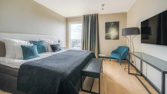 A bed with teal and gray pillows is neatly made; beside it, a chair sits under a large window with curtains, illuminated by a floor lamp. A wall-mounted TV completes the bedroom setting.