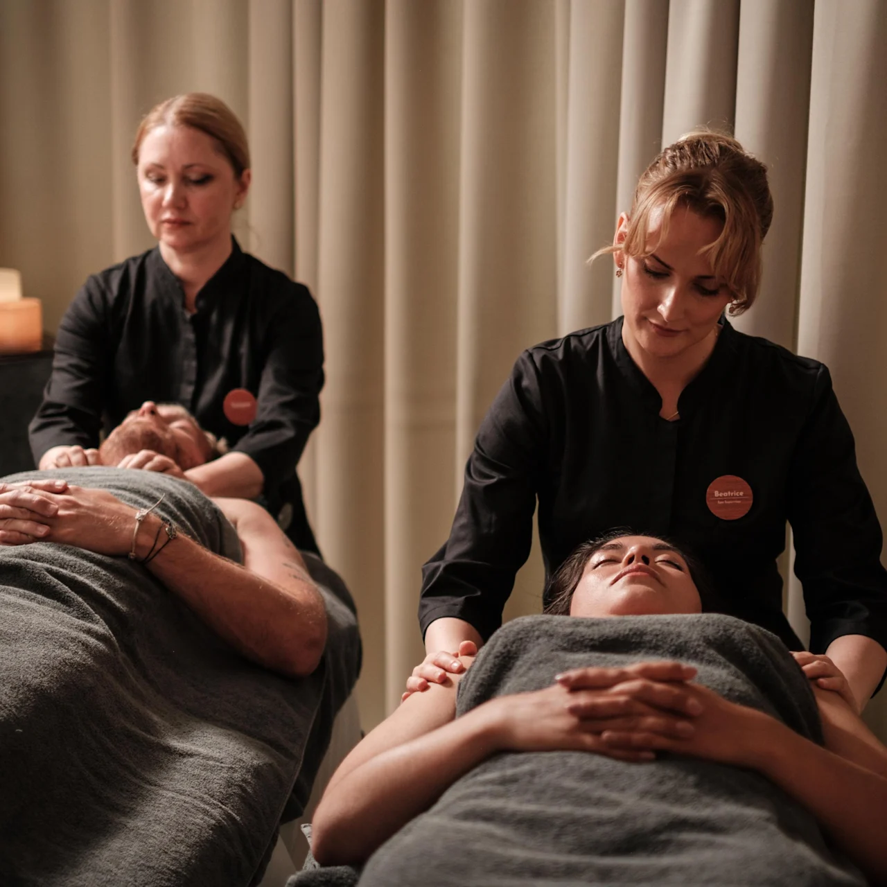 Två personer ligger under grå filtar och får massage av massageterapeuter i ett svagt upplyst rum med ljus och skapar en avkopplande atmosfär.