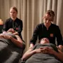 Två personer ligger under grå filtar och får massage av massageterapeuter i ett svagt upplyst rum med ljus och skapar en avkopplande atmosfär.