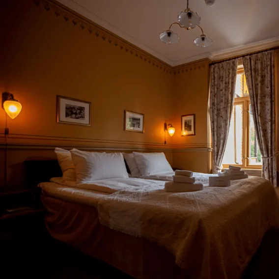 En seng med hvitt sengetøy og pent brettede håndklær er i et koselig, varmt opplyst rom med beige vegger, dekorative vegglamper, innrammede bilder og et vindu med mønstrede gardiner.