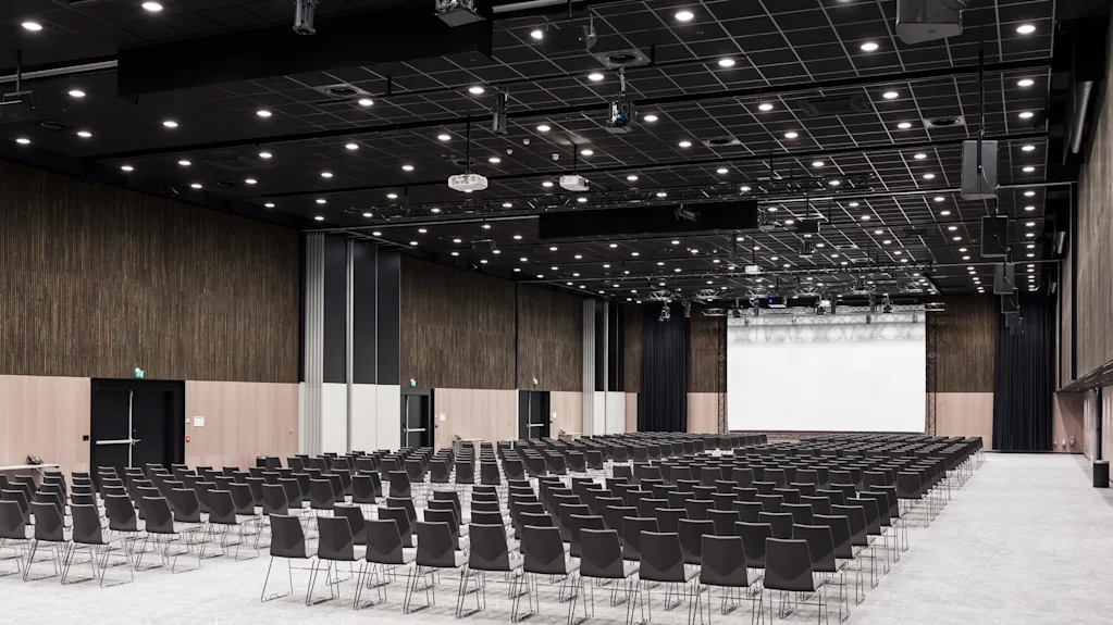 Rader av tomma svarta stolar vetter mot en stor projektionsskärm i en rymlig, modern konferenssal med högt i tak och mönstrat golv.