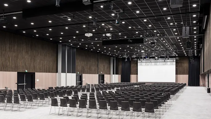 Rows of empty black chairs face a large projection screen in a spacious, modern conference hall with high ceilings and patterned floor. 