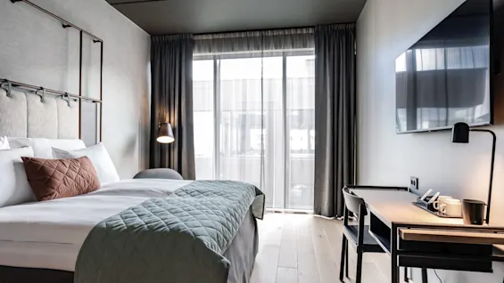 En prydligt bäddad säng med texturerade kuddar och vadderat täcke står i ett modernt hotellrum, bredvid en väggmonterad TV och ett skrivbord med en lampa och kaffekoppar.
