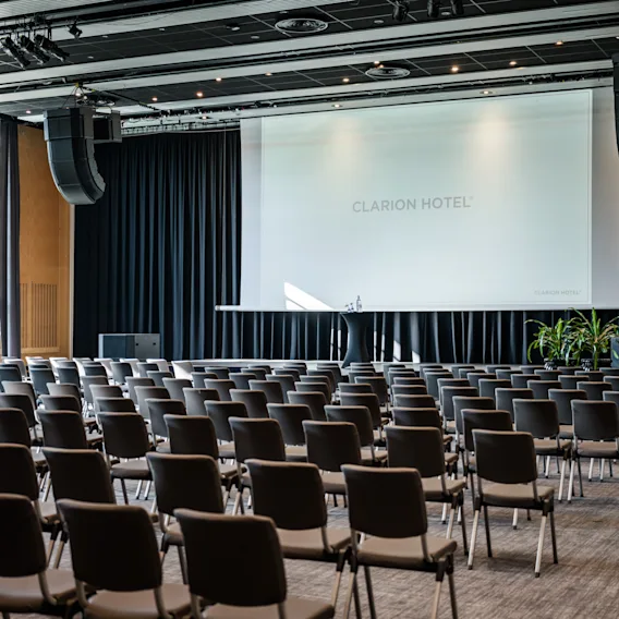 A spacious conference room with rows of chairs facing a large projection screen, set up for a presentation or event.