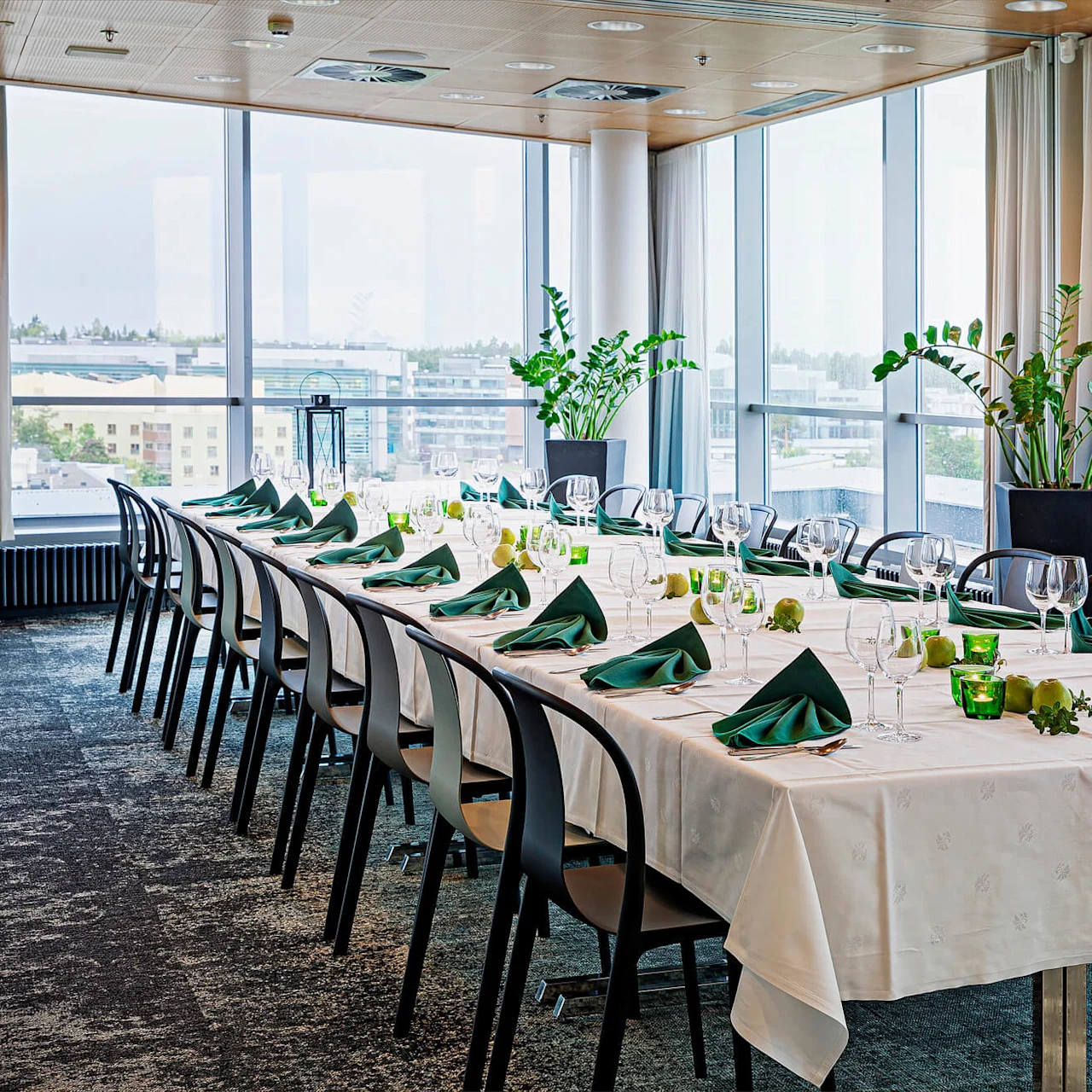 A long dining table set with white cloth, green napkins, glassware, and decorative apples is surrounded by black chairs in a bright room with large windows and potted plants.