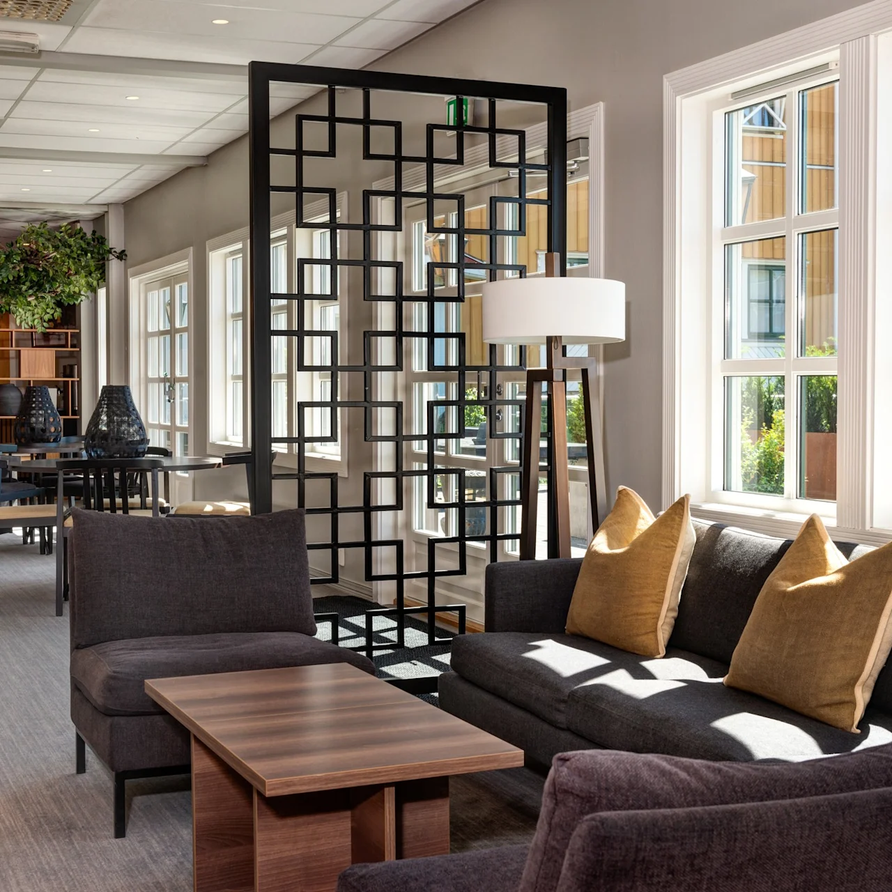 Dark gray sofas with mustard pillows are arranged around a wooden coffee table. Modern black room divider stands nearby. Sunlight streams through large windows, illuminating the room with plants.