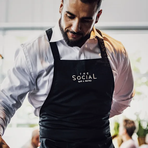 En servitør med svart forklé merket "The Social Bar & Bistro" som ser oppmerksomt ned.