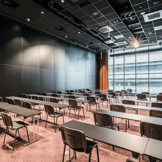 Rader med svarte skrivebord og stoler arrangert i et tomt konferanserom med høyt tak og store vinduer med utsikt over en bygning utenfor, pyntet med oransje gardiner.