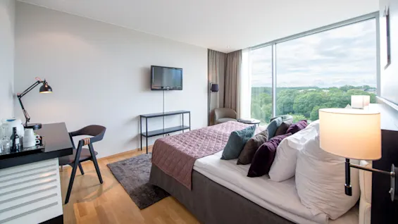 Et moderne hotellrom har en ryddig seng med fargerike puter, et skrivebord med lampe, en veggmontert TV og store vinduer med utsikt over frodig natur utenfor.