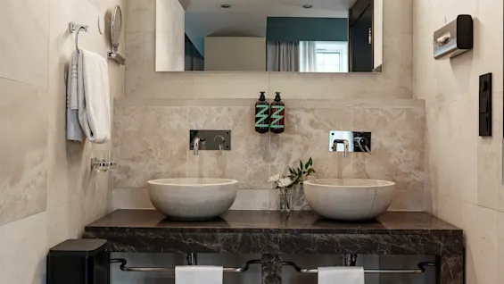 Two round stone sinks with running water sit on a dark countertop beneath a large mirror. Towels and soap dispensers accompany them in a modern bathroom setting.