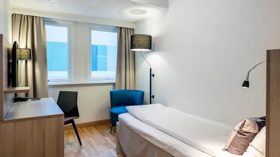 A neatly made single bed stands against the right wall, flanked by a floor lamp and small desk with a chair, in a cozy, minimalist room with wood flooring.