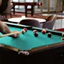 A person holds a pool cue, aiming at billiard balls on a green felt table in a lounge with chairs, a round table, and glass doors in the background.