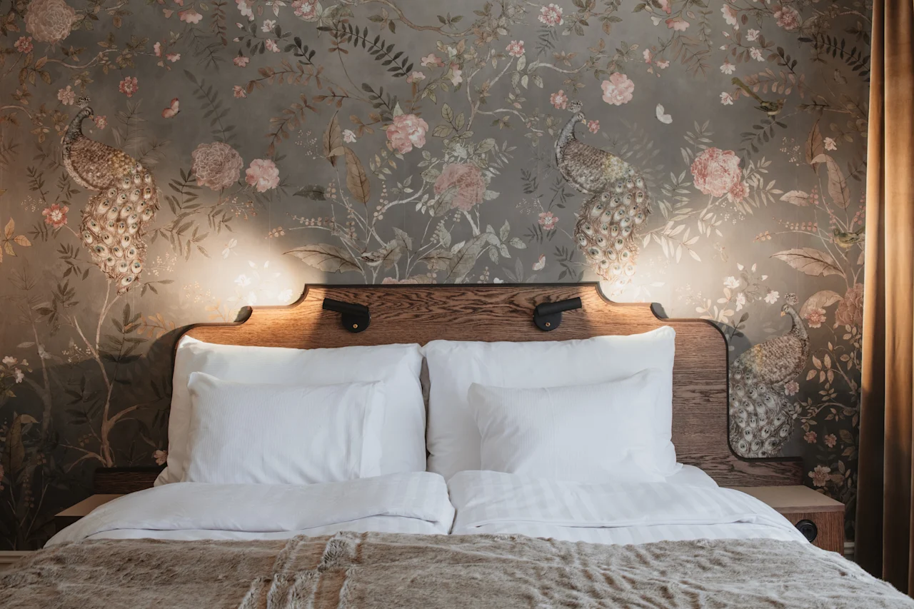 Home Hotel Baltzar - Deluxe Double. A cozy bed with a wooden headboard and elegant peacock and floral wallpaper.
