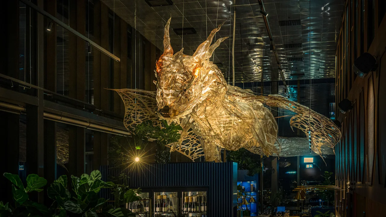 En gjennomskinnelig, opplyst drageskulptur henger i et moderne rom med høyt tak og store vinduer, med planter under.