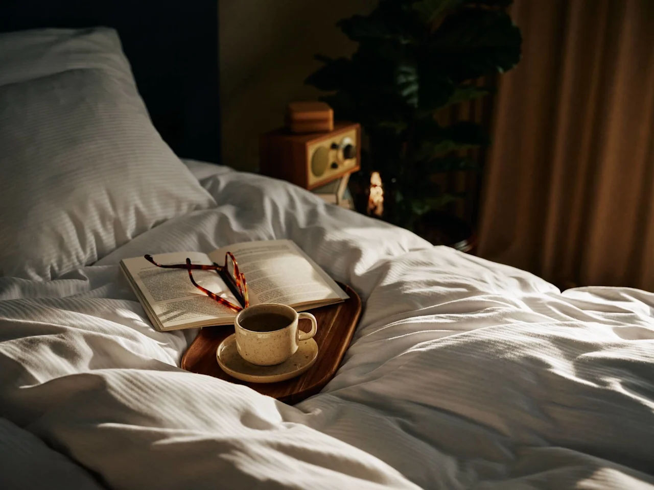 En bog og briller hviler på en sengbakke med en rygende kaffekop, omgivet af hvidt sengetøj i et hyggeligt soveværelse, blødt oplyst af naturligt lys.
