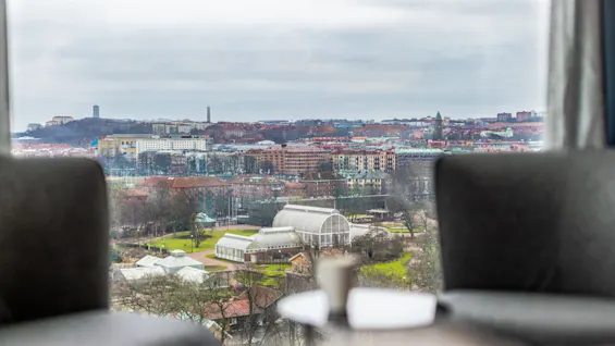 A distant view of a greenhouse in a park, surrounded by urban buildings under a cloudy sky, framed by two blurred chairs.