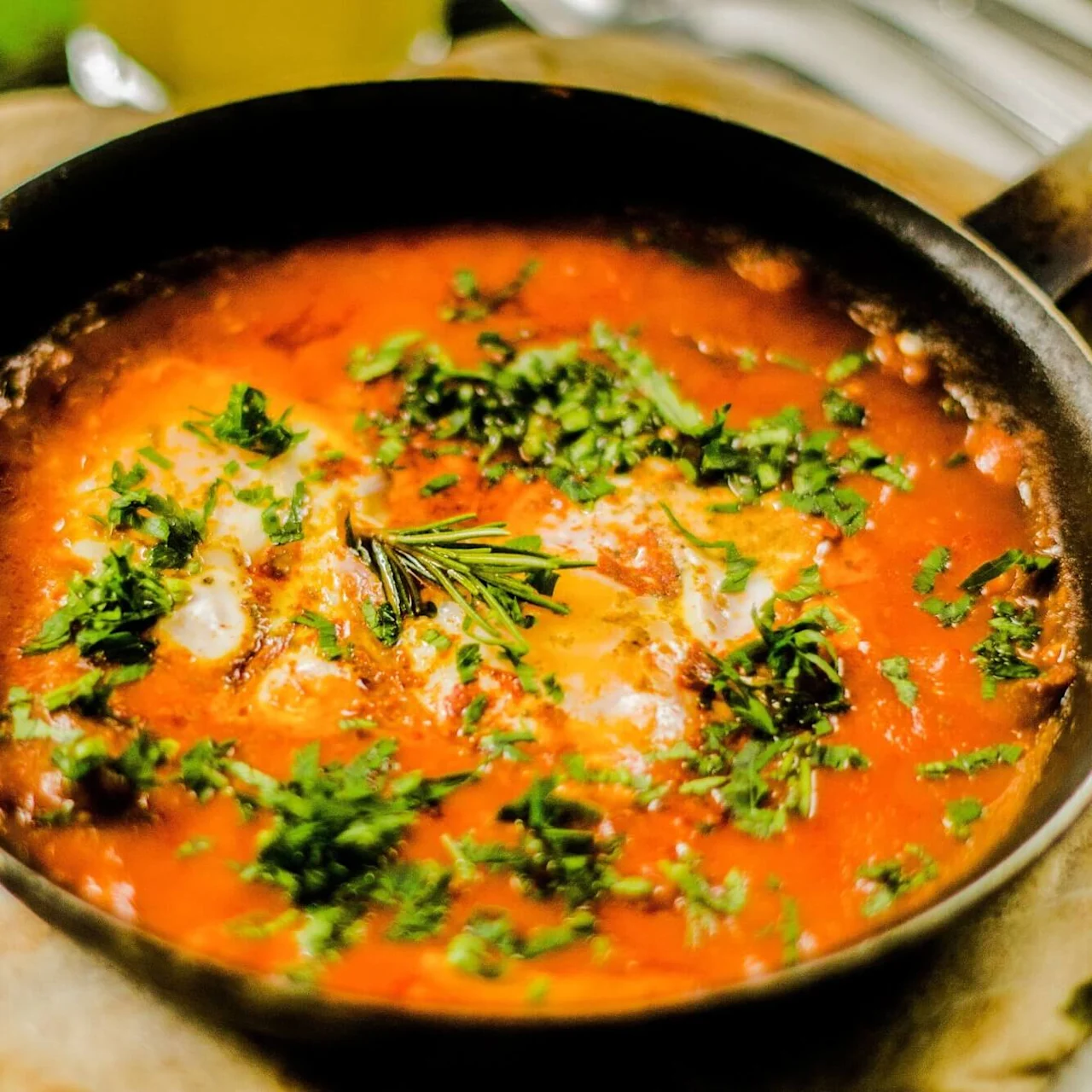 En stekepanne med tomatbasert rett med urter på siden og flatt brød på et bord.