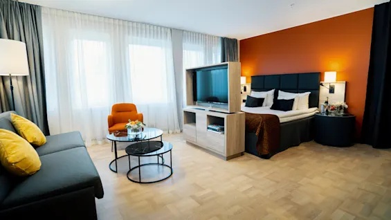 Ett modernt hotellrum med en säng med vita lakan, en orange fåtölj och en grå soffa. Ett trävitrinskåp delar upp rummet. Stora fönster lyser upp rummet.