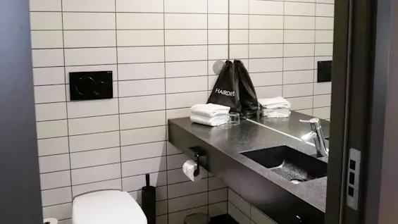 Vask reflekteres i et baderomsspeil, flankert av en hengende hårfønerpose og foldede håndklær; toalett under hylle med håndklær, med lyse lamper over.