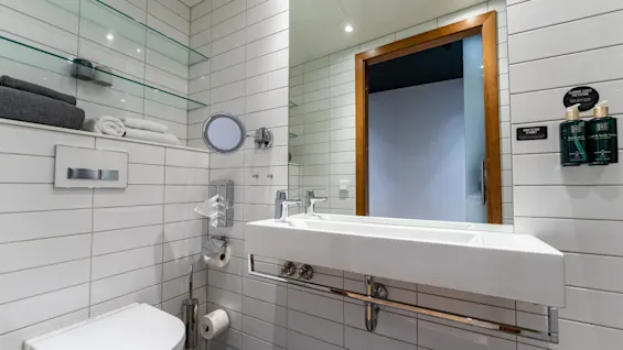 Et moderne badeværelse med en hvid vask og toilet, glashylder med håndklæder, og sæbedispensere. Væghængt spejl over vasken og dør som fører til et andet rum. Tekst: "CLAIM YOUR LINKS THE FUTURE. WANT TO TAKE THEM WITH YOU?"