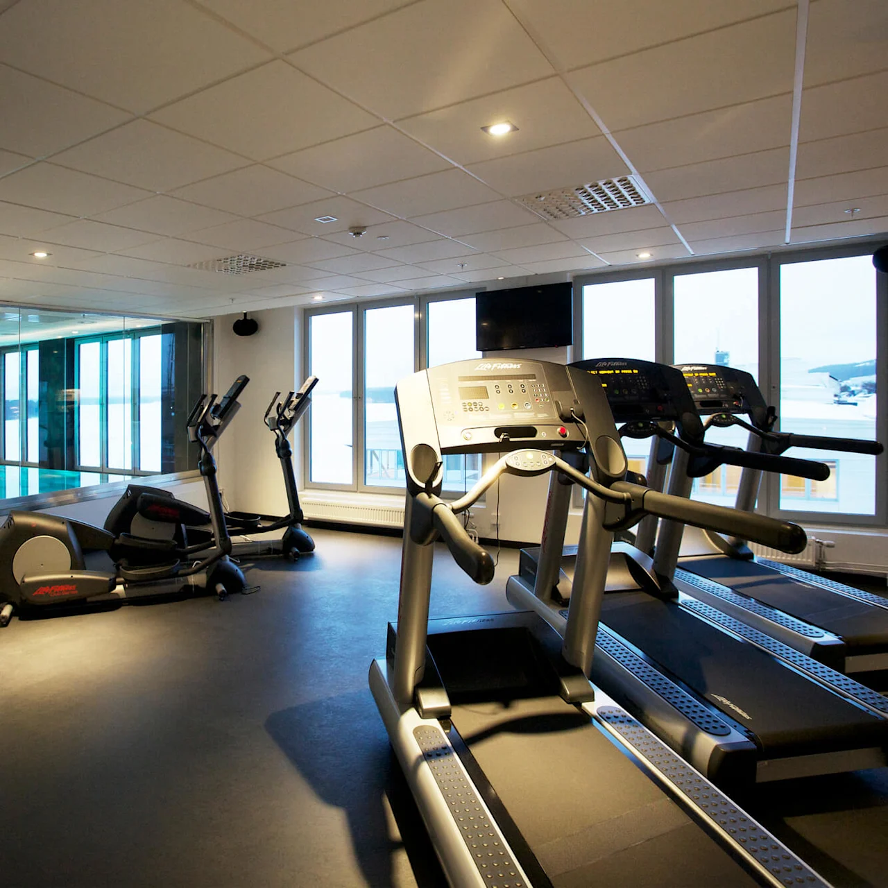 Löpband står stilla i ett modernt gym och vetter mot stora fönster som vetter mot en stadsbild. Crosstrainers och träningsutrustning flankerar dem under ett tak med jämnt fördelade lampor.