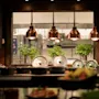 A warm and inviting food service area with a glimpse into a busy kitchen, featuring copper lamps and fresh plants.