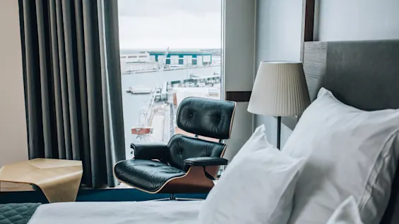 En luksuriøs sort lænestol står vendt mod et vindue med udsigt over en havn. Hvide puder ligger på en seng ved siden af en lampe i et stilfuldt hotelværelse.