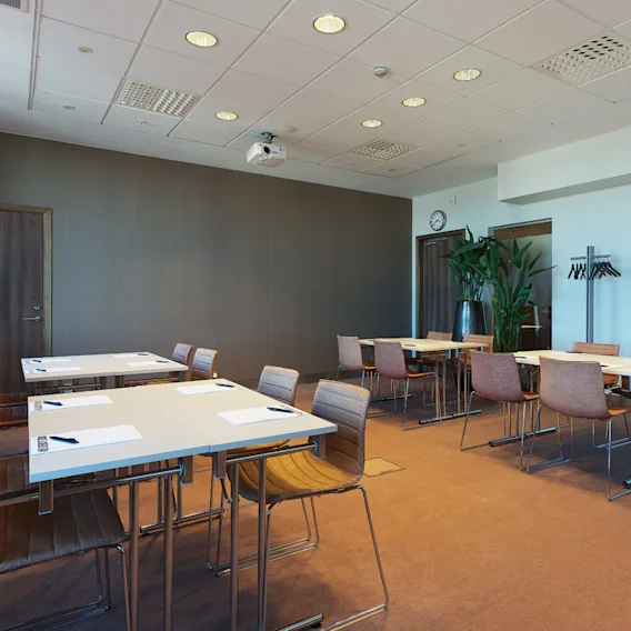 Bord och stolar är arrangerade i ett konferensrum med anteckningsblock och pennor på borden. En projektor hänger från taket och en växt dekorerar ett hörn.