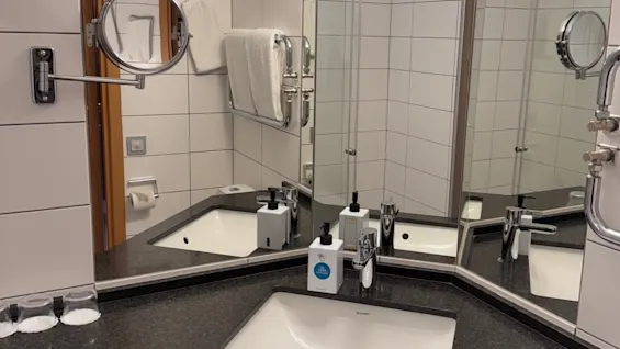 A triangular bathroom sink is set in a corner with black countertop, surrounded by mirrors. Soap dispensers and glasses are placed near the faucet, with towels on a wall rack.