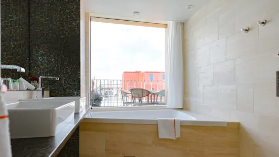 A modern bathroom with a large bathtub below a window, surrounded by beige tiles. Outside, wicker chairs and a table sit on a balcony, overlooking a red building.