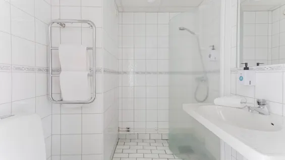 A clean, white-tiled bathroom featuring a shower with a glass partition, a toilet, and a sink with a mirror.