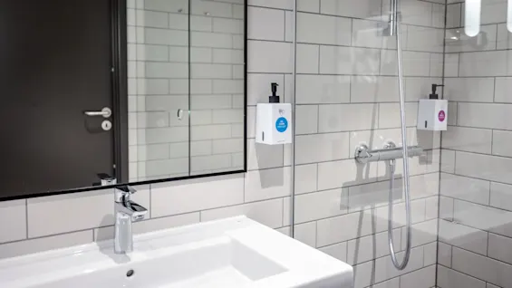 Et hvidt badeværelse har en elegant vask og vandhane under et spejl og en grå dør. Væghængte sæbedispensere lyder "Body Wash" og "Hair & Body Shampoo" ved en bruser.