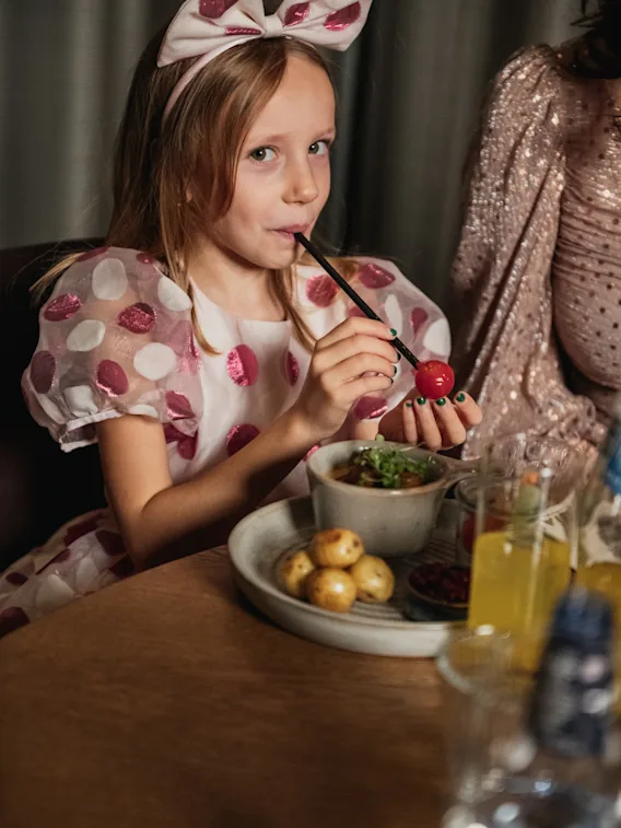 En ung jente har på seg en prikkete kjole med sløyfe og drikker gjennom et sugerør. Hun sitter ved et bord med en tallerken små poteter og salat.