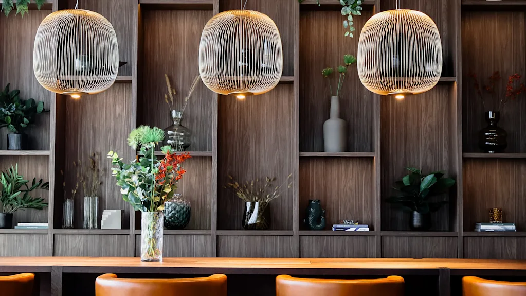Suspended lights hang above a wooden table adorned with a vase of flowers. Wooden shelves in the background hold various decorative items and plants, creating a cozy atmosphere.