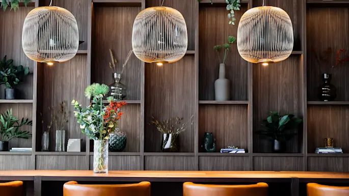 Lamper henger over et trebord med en vase med blomster. Bakgrunnen har trehyller med dekorative gjenstander og planter, og skaper en koselig atmosfære.