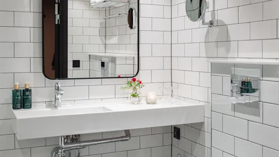 Moderne badeværelse med en hvid vask under et rektangulært spejl, flisebelagte vægge og pænt foldede håndklæder på et metalstativ. Et toilet og små dekorative genstande tilføjer funktionalitet og elegance.