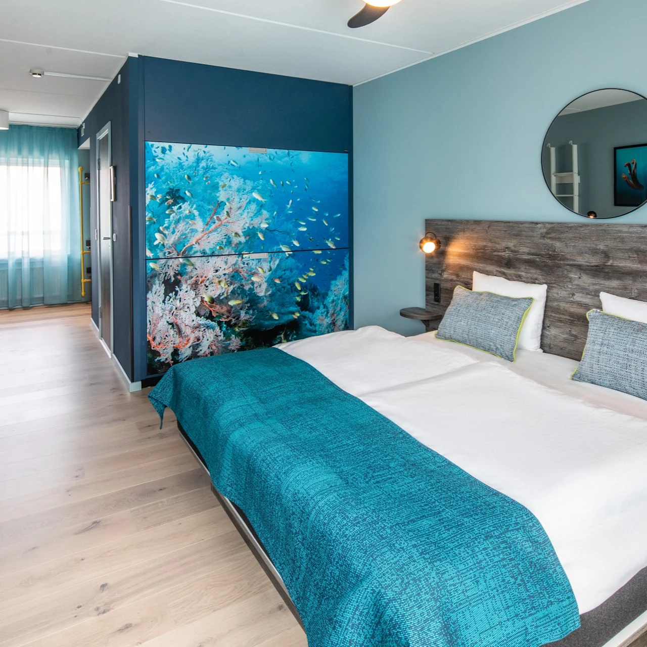 A neatly made bed with turquoise bedding rests under a circular mirror. Wall art depicting an underwater scene adorns a light blue hotel room, complemented by a desk and chair.
