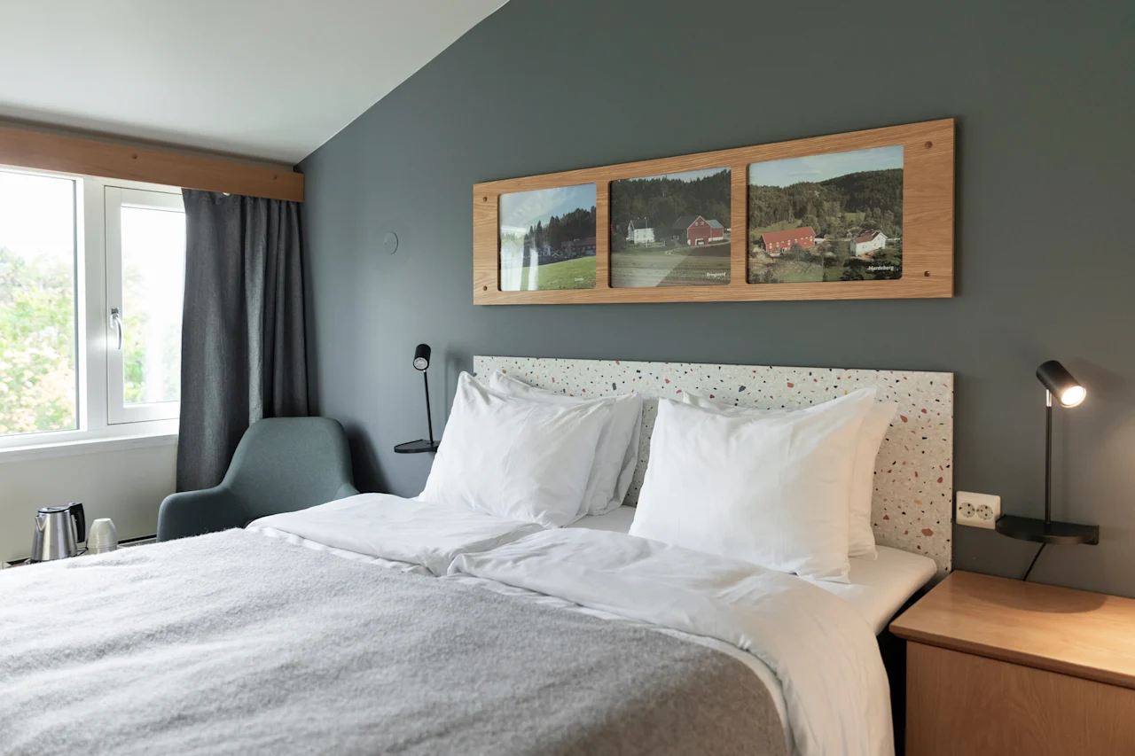 A cozy hotel room with a comfortable bed, framed landscape art, and a window offering a view of nature.