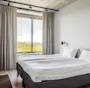 En pent oppredd dobbeltseng med hvitt sengetøy står ved et stort vindu med lys grå gardiner, i et minimalistisk rom med tregulv og en veggmontert lampe.