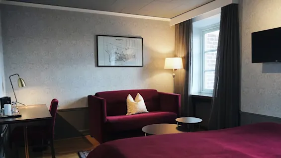 En rød seng i et koselig rom ved siden av en matchende sofa og to runde bord; et skrivebord, lampe og vindu forbedrer omgivelsene. Et innrammet kart henger på veggen.