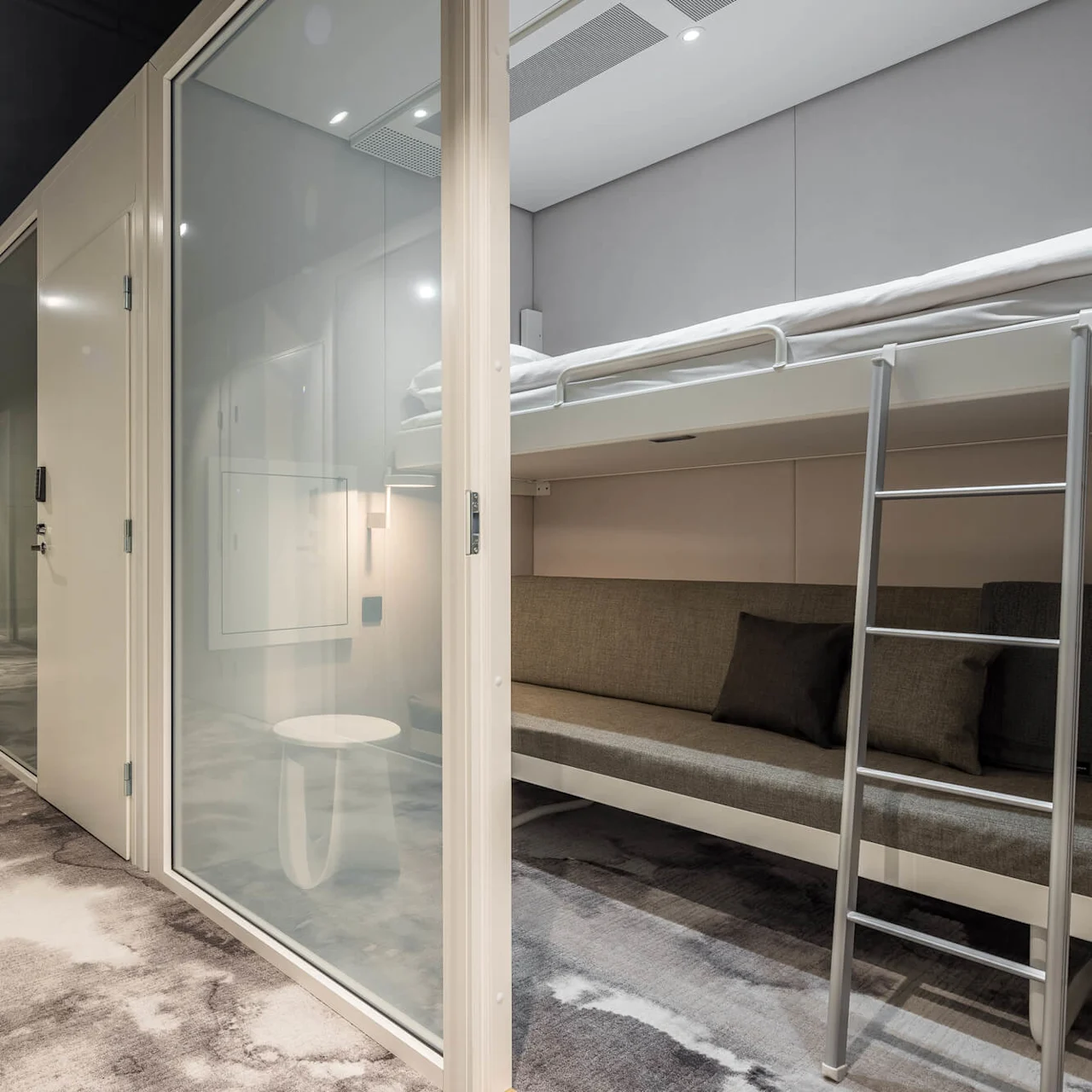 Kompakta sovkapslar med våningssängar i en modern miljö. Hallen har mönstrad matta, upplyst av taklampor, med dimmig skogstapet i slutet.