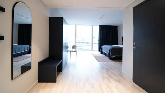 A sleek modern bedroom features a bed with dark bedding, a large window, light wooden floor, and a mirror reflecting part of the bed and room. 