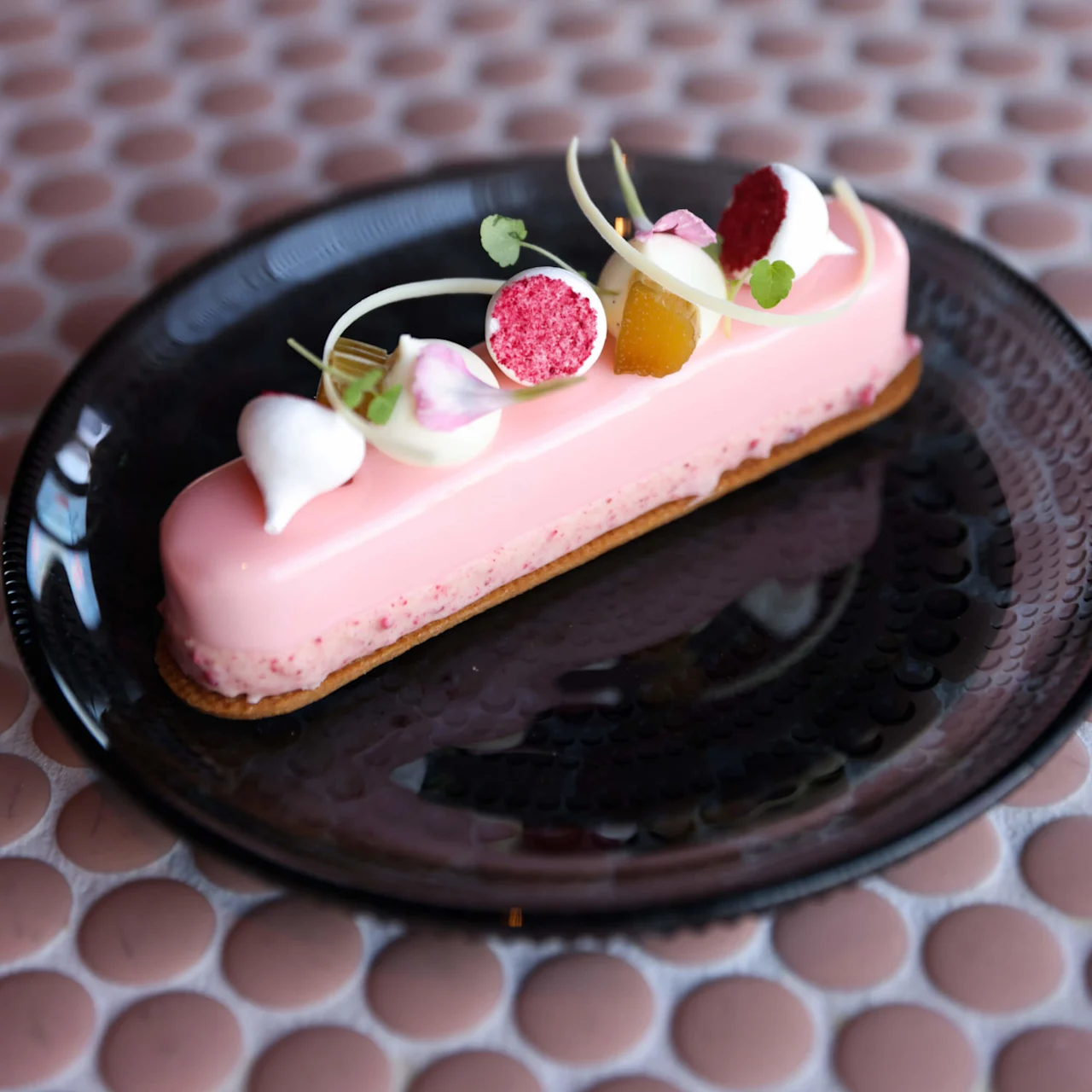 Elegant rosa dessert ligger på en svart tallrik, garnerad med krämdroppar och ätbara blommor, placerad på ett bord med cirkelmönster.