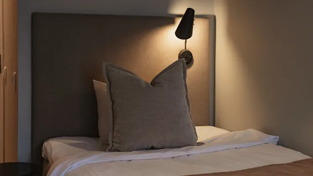 En bäddad säng har en stor grå kudde mot en vadderad sänggavel, belyst av en vägglampa ovanför. Rumets mjuka belysning skapar en varm, inbjudande atmosfär.