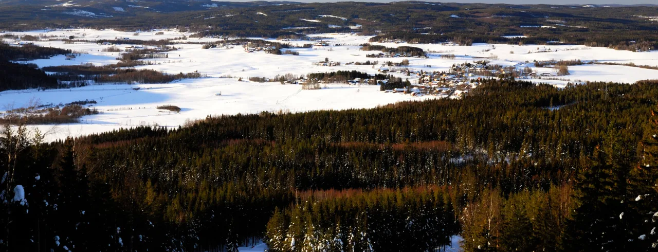 En snötäckt landskap med en by, skogar och kullar under en klarblå himmel.