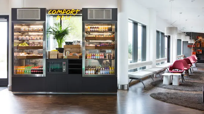 A vending area displays snacks, drinks, and a microwave under a "Comfort Deli" sign, set in a bright, modern lounge with chairs and large windows.