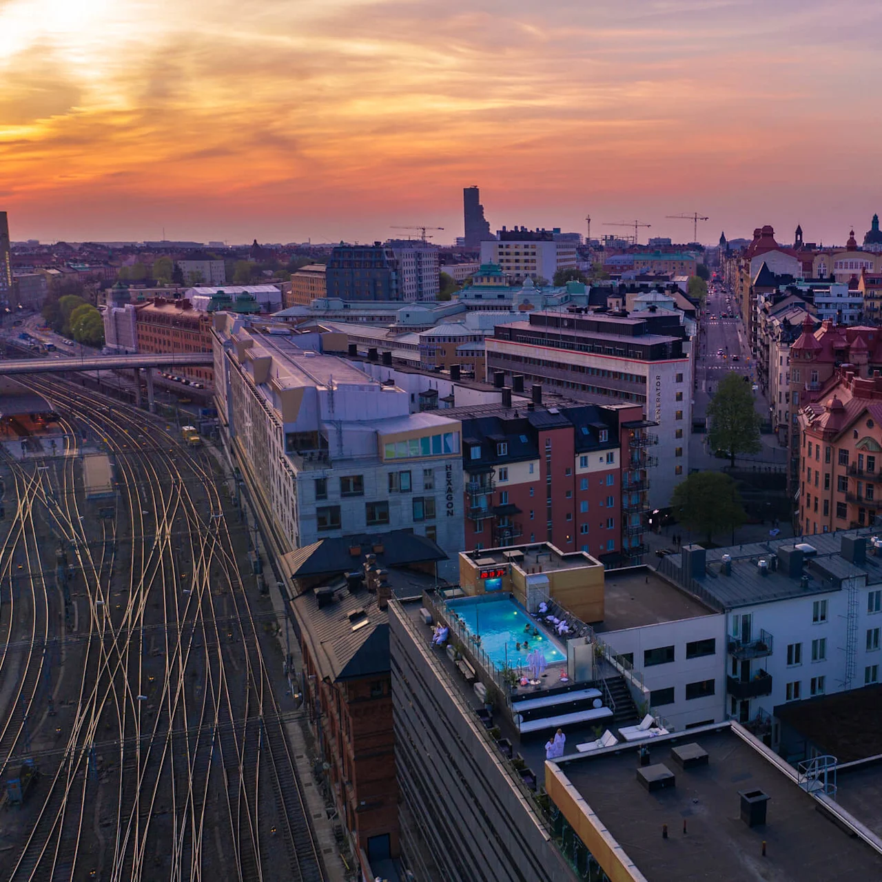 Takpool omgiven av människor, med utsikt över en vidsträckt stadssilhuett och järnvägsspår i solnedgångsljus.