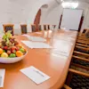 Bowl of assorted fruits sits centered on a long conference table, surrounded by chairs, with a notepad and pen at each seat; glasses and candles are arranged near a screen.