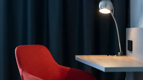 En röd stoppad stol står bredvid ett skrivbord i trä med en tänd metallisk lampa, mot mörka gardiner.