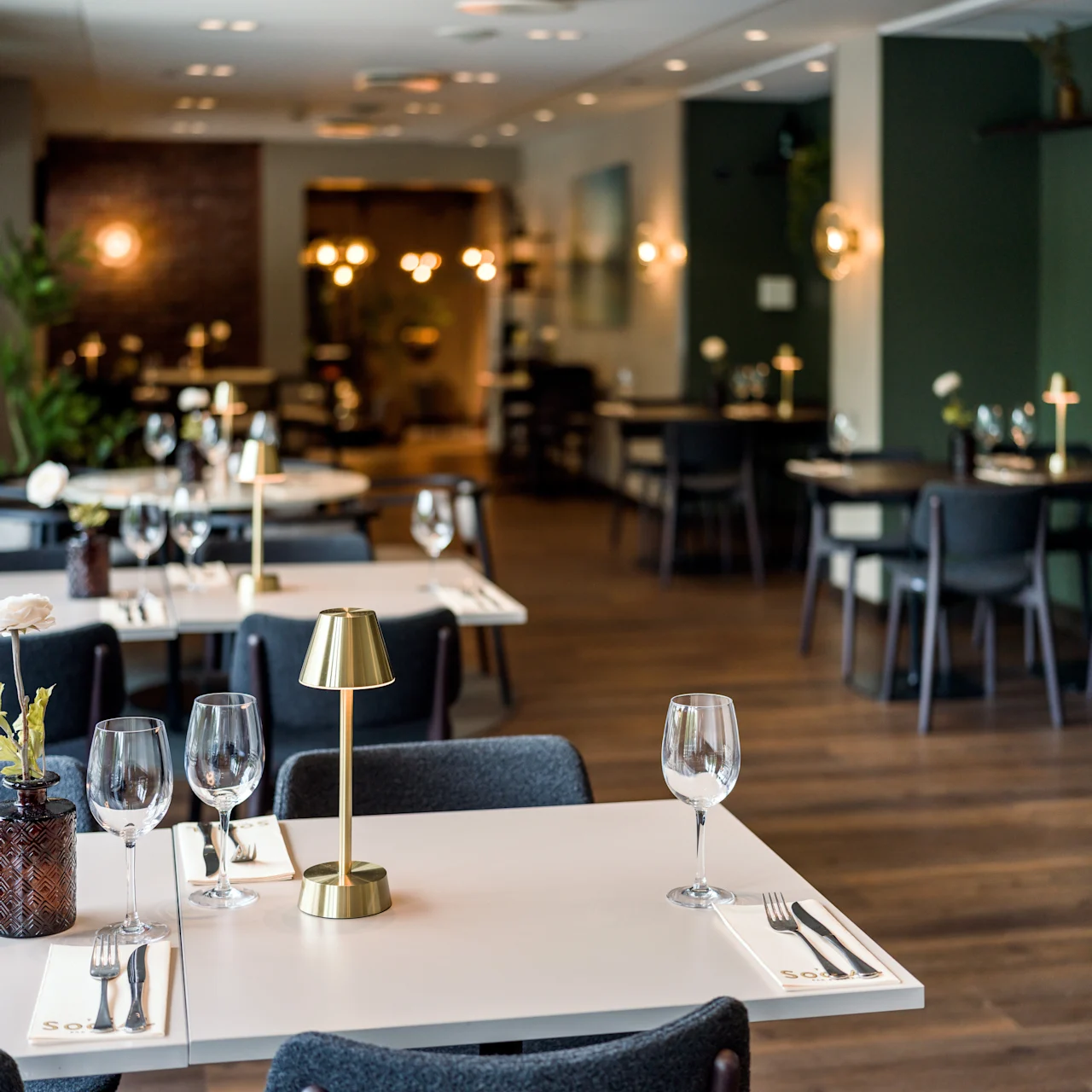 Tastefully set dining tables with wine glasses and small lamps in an elegant restaurant, featuring ambient lighting and dark, modern decor creating a warm, inviting atmosphere.